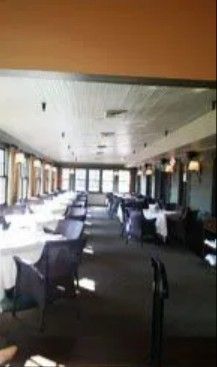 An empty restaurant dining area with rows of tables, white tablecloths, and dark chairs under a long, white-paneled ceiling.