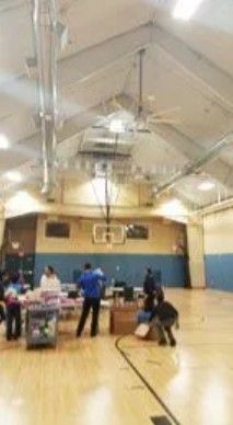People preparing tables and supplies in a gymnasium with a basketball hoop on the wall.