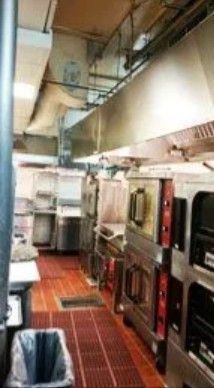 A commercial kitchen features stainless steel ovens under a large exhaust hood, with a trash can and red floor mats.