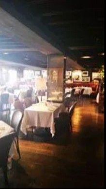 A dimly lit restaurant interior with tables covered in white tablecloths, dark wooden floors, and a low, beamed ceiling.