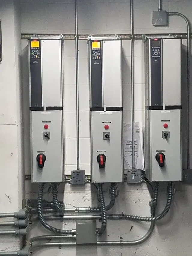 Three industrial motor control drives mounted on a white brick wall, featuring silver panels and black rotary switches.