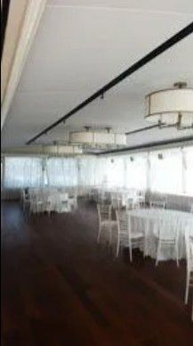 A bright, spacious event hall featuring round tables with white tablecloths, chairs, and modern drum pendant lights.