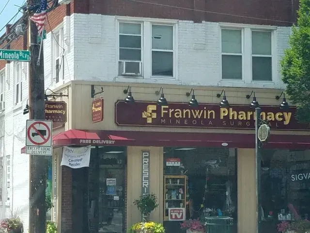 The storefront of Franwin Pharmacy on a corner with a red awning and white brick upper level under a bright sky.