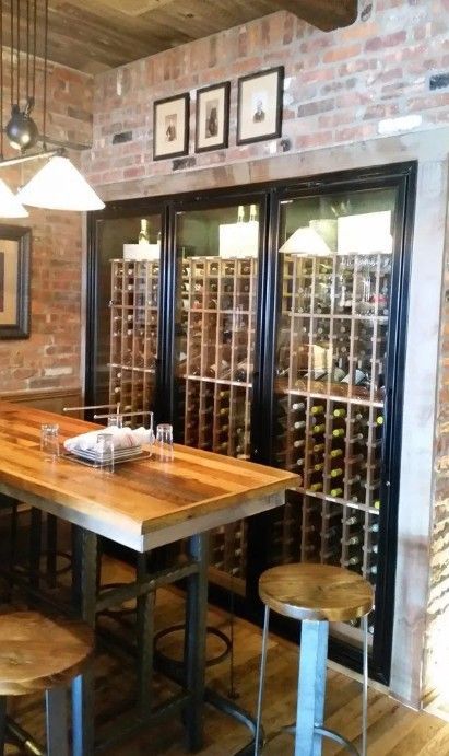 A rustic indoor bar area with a wooden high-top table, stools, and a large triple-door glass wine cellar against brick.