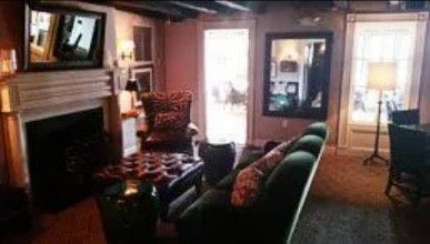 A cozy living room with a fireplace, a green sofa, a leather armchair, a patterned ottoman, and two large mirrors.