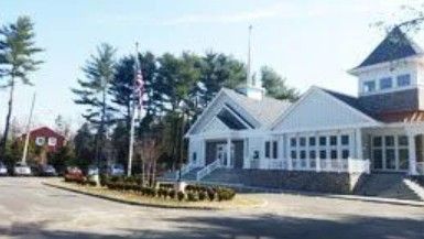 A white building with a steeple and large windows, featuring a flag pole and circular driveway on a sunny day.