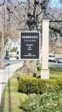 A black sign for Hendrick's Tavern, featuring restaurant, bar, and event details, on a post near a road and green hedge.