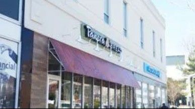 A Panera Bread restaurant with a maroon awning on a light-colored building in a downtown area.