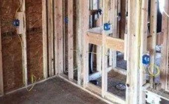 An interior view of a room under construction, showing wooden wall studs, blue electrical boxes, and wiring.