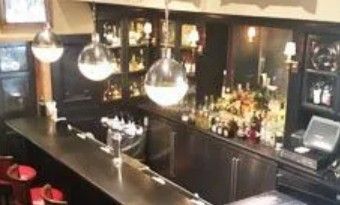 A high-angle view of a dark wooden bar counter with stools, glass globe pendant lights, and backlit shelves of bottles.
