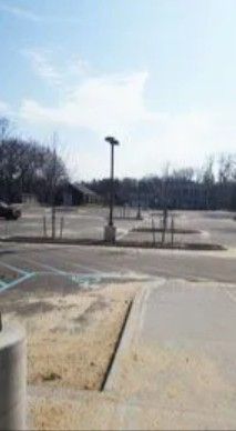 An outdoor parking lot with a tall light pole, some young trees, and a small building in the distance under a blue sky.