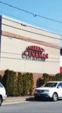 The Merrick Cinemas building exterior, featuring red lettering on a beige wall above a row of shrubs and parked cars.