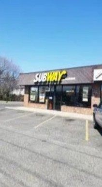 A Subway restaurant exterior with a yellow sign above a glass storefront, set in a parking lot under a clear blue sky.