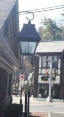 A hanging black metal lantern fixed to a building corner overlooking a street with a Tudor-style storefront in the distance.