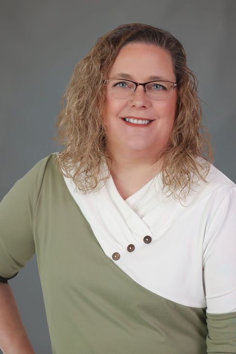 Kim Ankrum with curly hair, glasses, and a smile wearing a green and white top, gray backdrop.