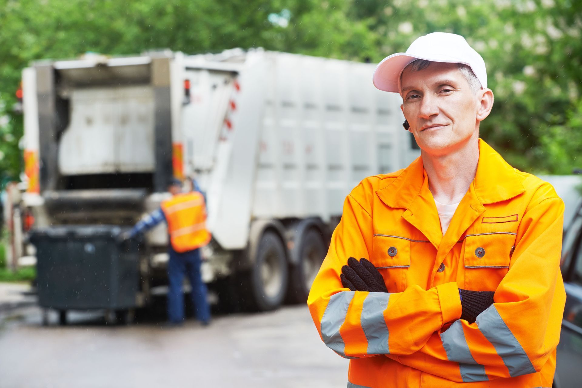 Garbage truck worker in orange uniform stands in front of a truck, arms crossed. Another worker loads a bin.