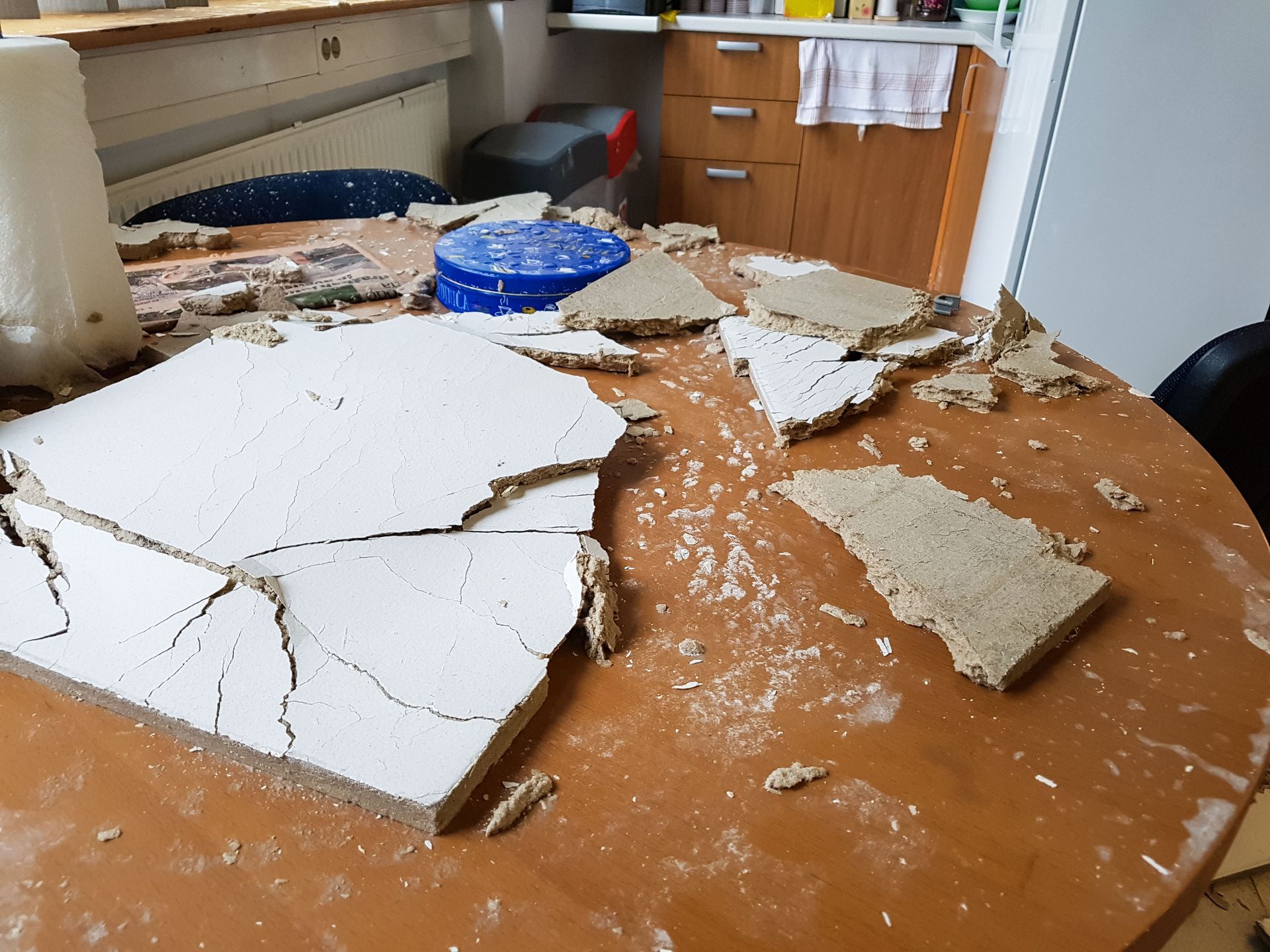 Broken drywall pieces on a wooden table, in a kitchen setting. White, beige, and brown colors are visible.