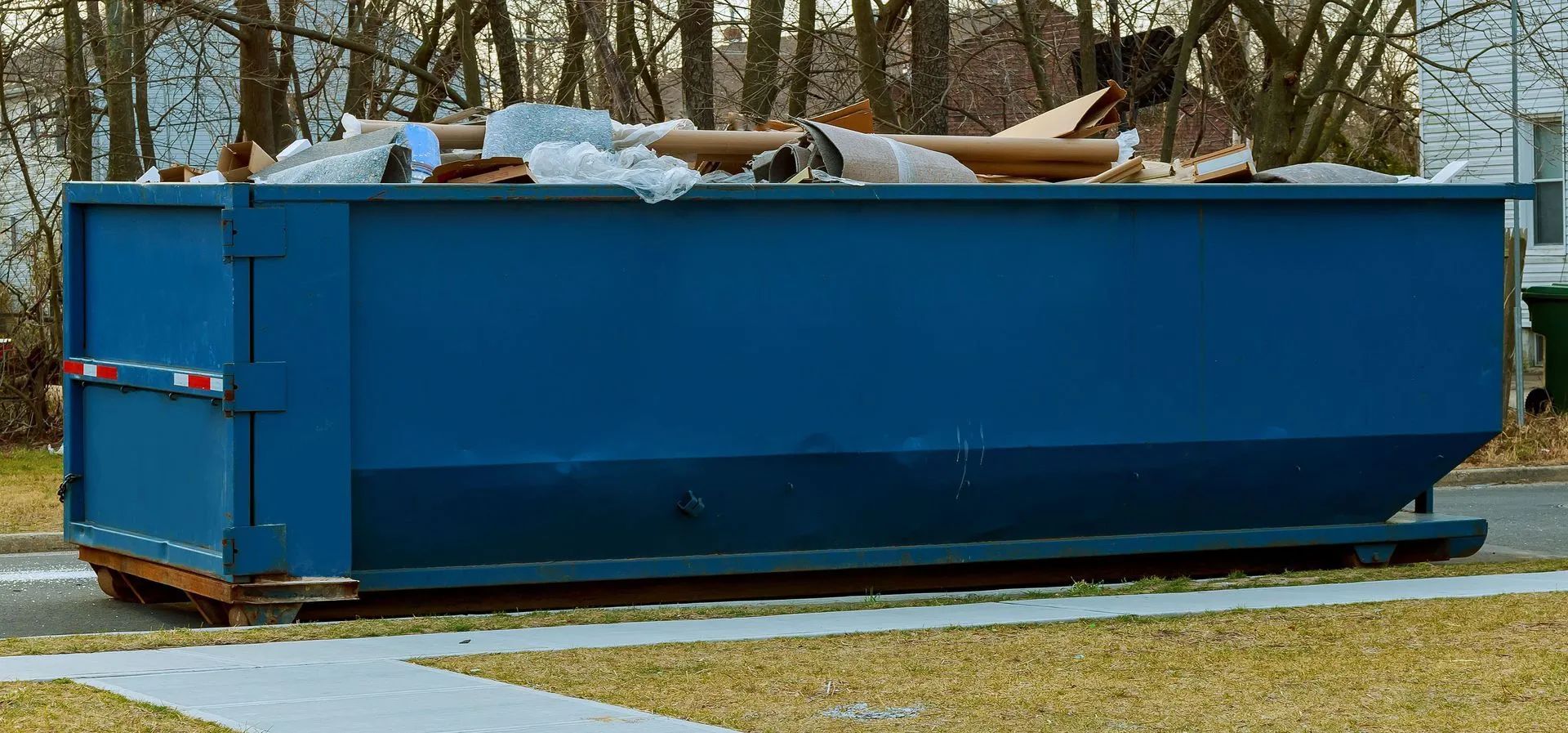 Blue dumpster filled with trash sits in a yard.