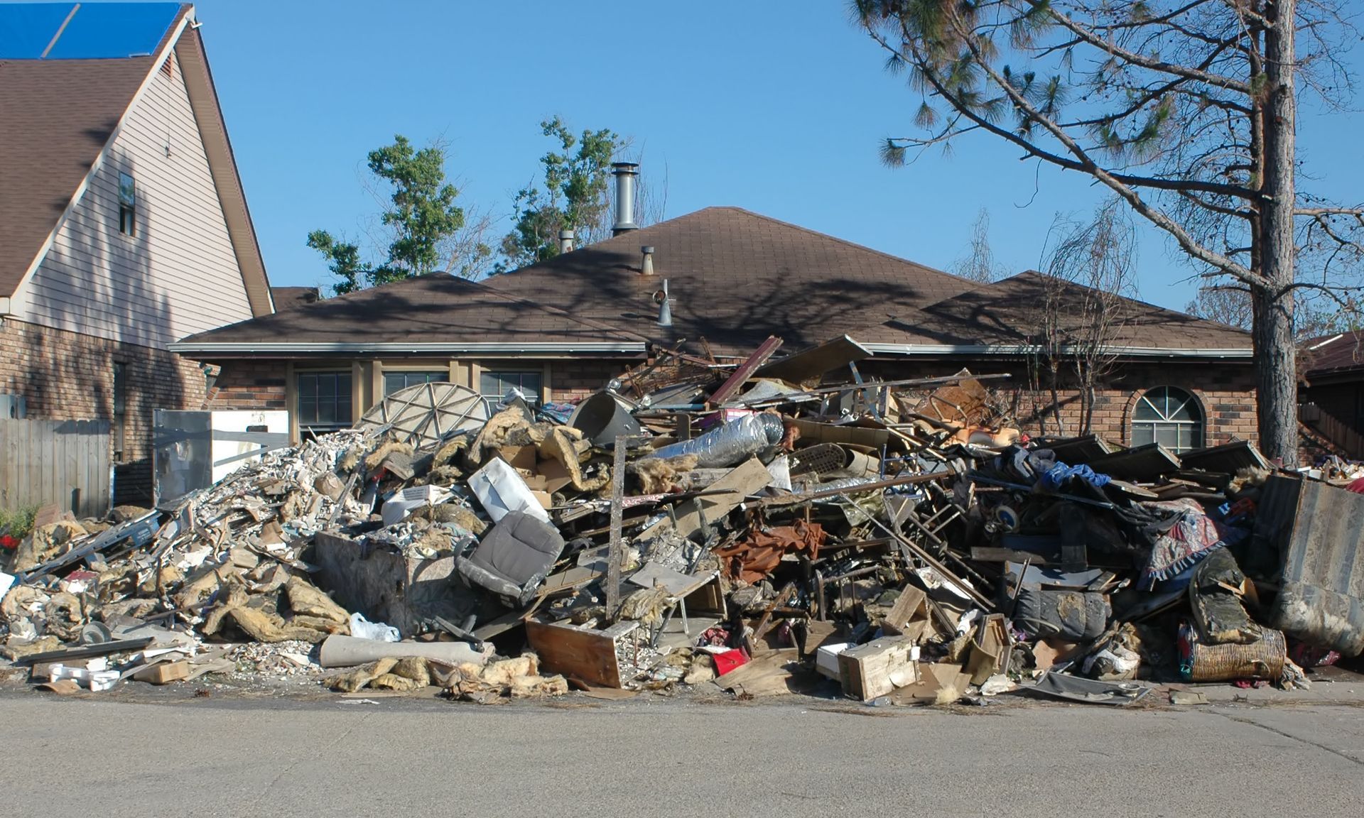 Pile of debris in front of a house after a disaster; bricks, furniture, and wood scattered.