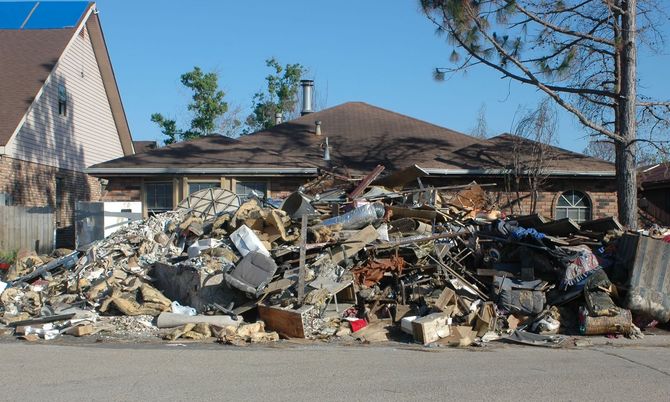 Pile of debris in front of a house after a disaster; bricks, furniture, and wood scattered.