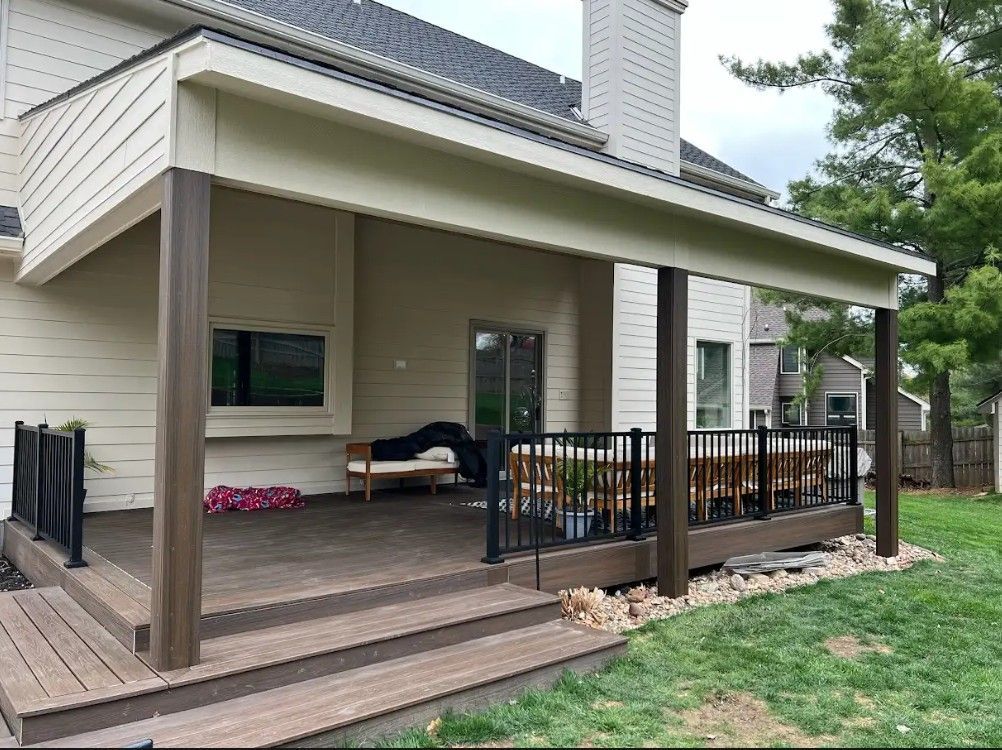 Covered deck with dark brown posts and railing attached to a beige house; view of yard.