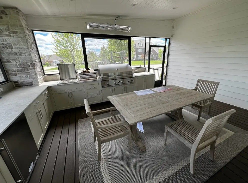 Outdoor kitchen with grill, table, chairs, and rug on a deck, enclosed by screen windows and a brick chimney.