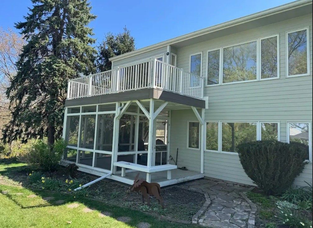 Two-story house with a screened porch, upper deck, and a dog on the grass. Green siding and a blue sky.