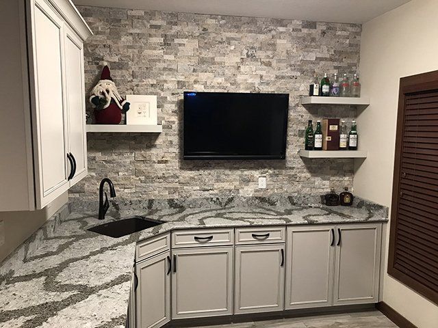 A kitchen with white cabinets , granite counter tops , a sink and a flat screen tv.