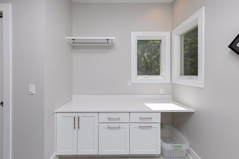 A laundry room with white cabinets , drawers , and two windows.