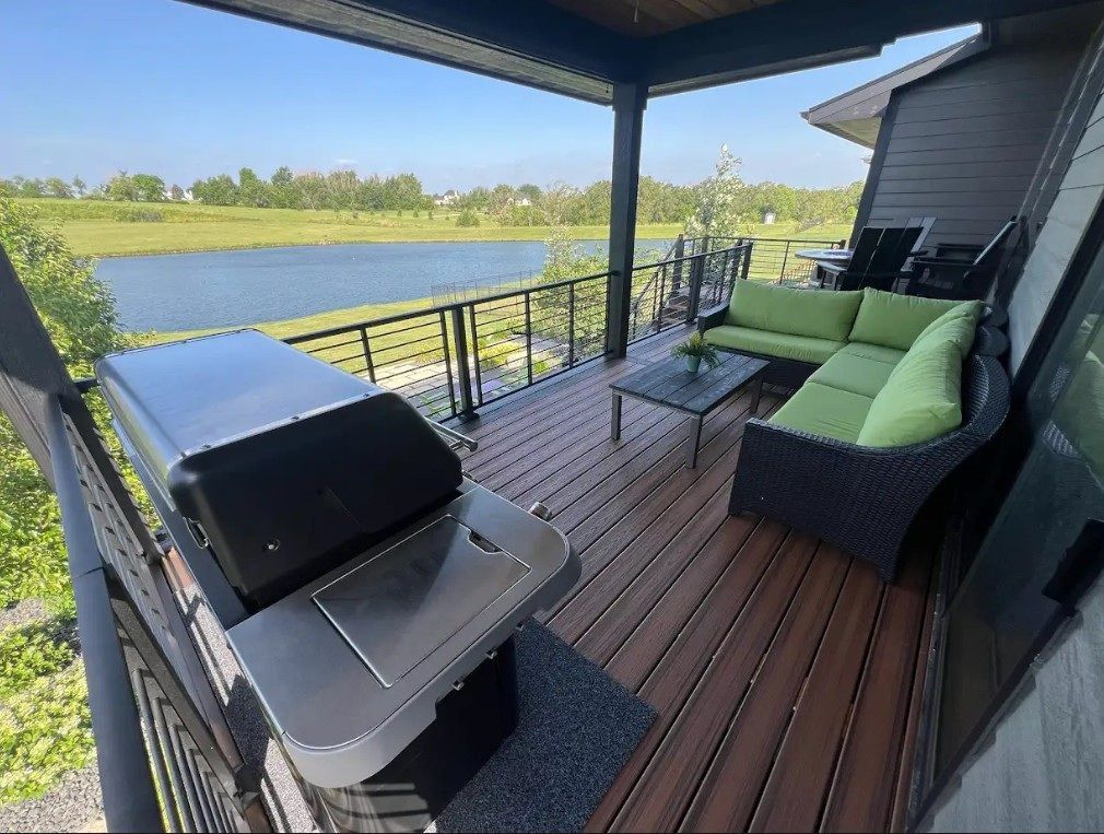 A deck with a grill, seating, overlooking a lake. Brown decking, black railing, and green cushions.