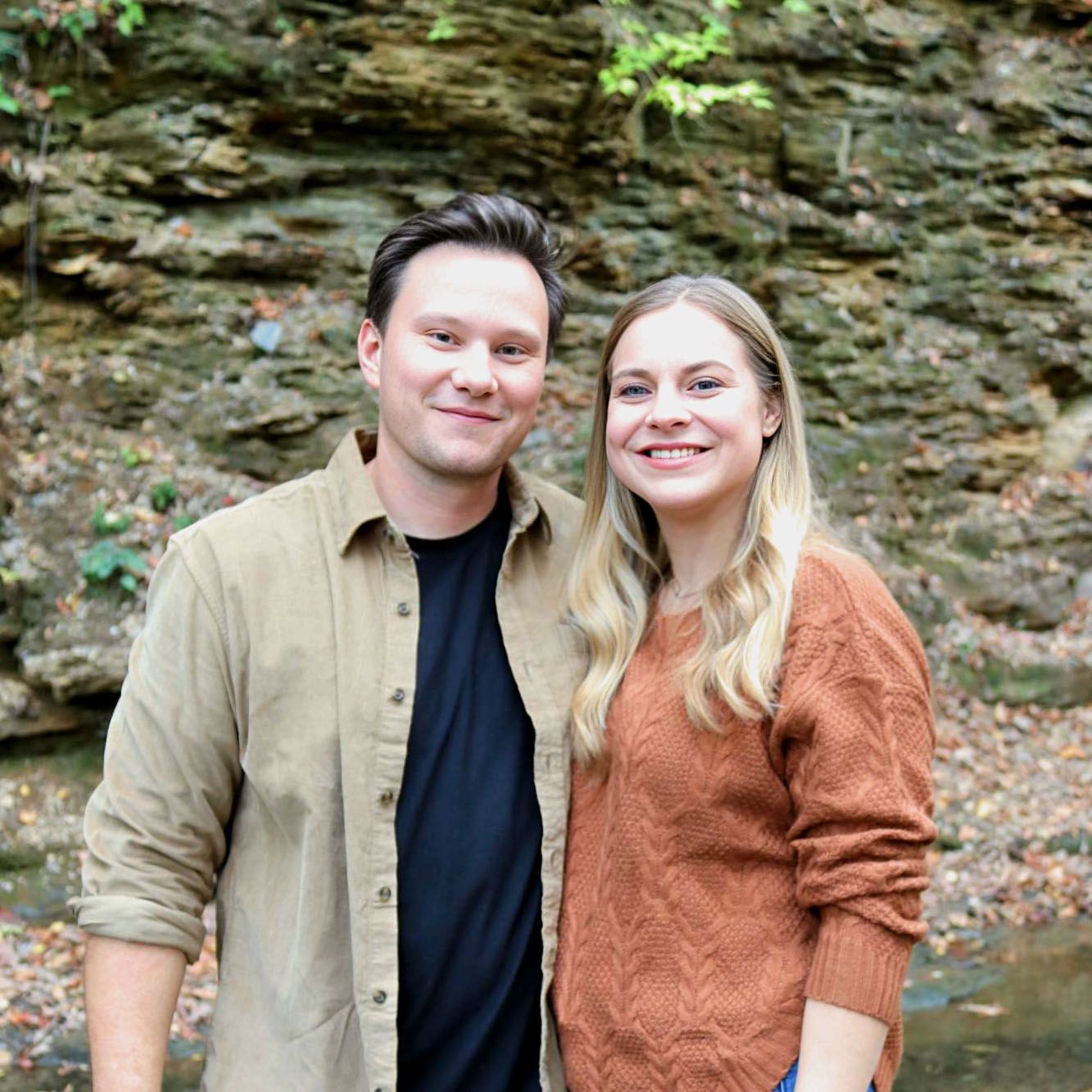 A smiling man and woman pose together outside near a rock wall. The man wears a tan shirt, and the woman wears an orange sweater.