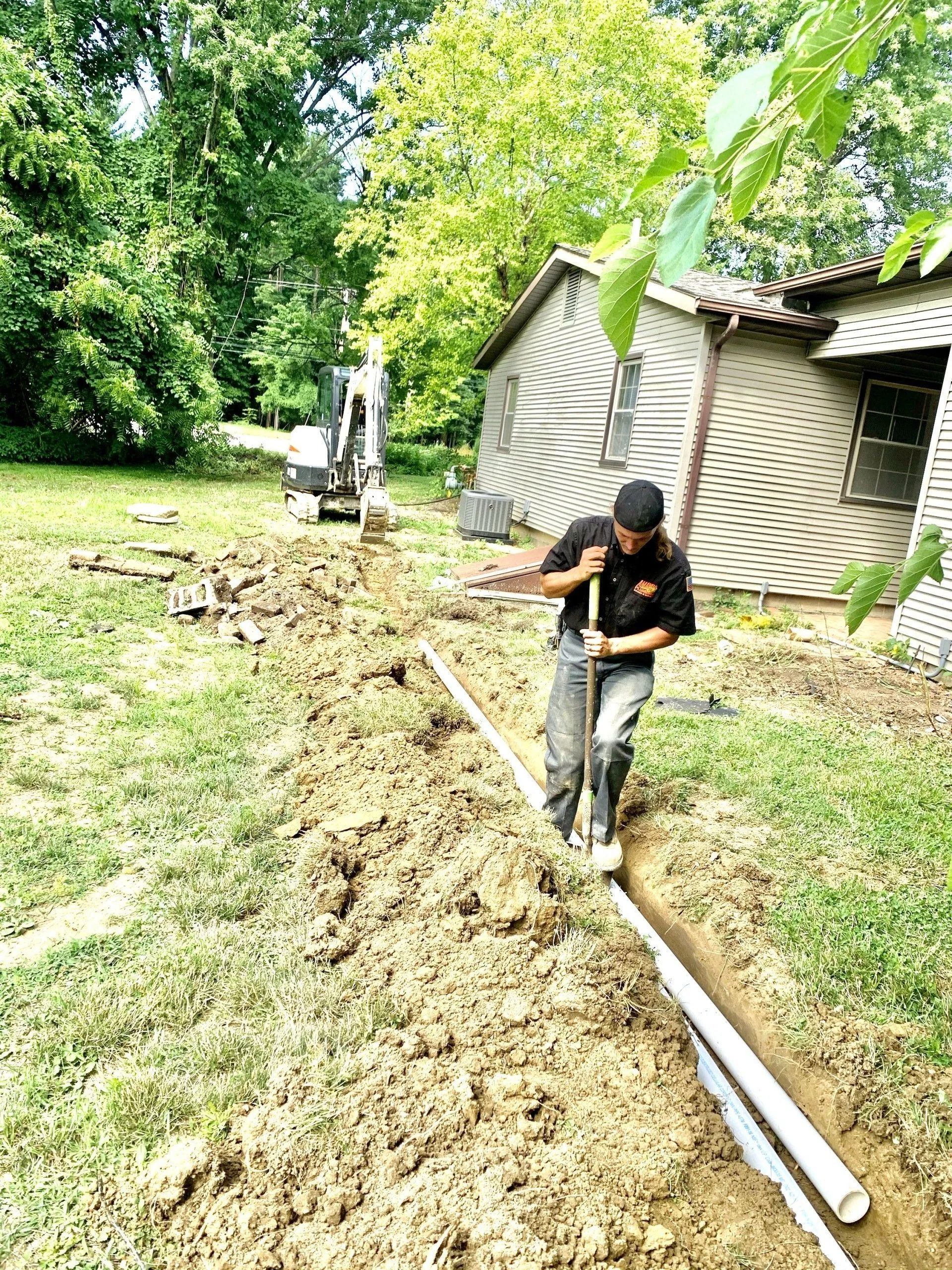 Man digging a trench, laying pipes near a building with a small excavator in the background.