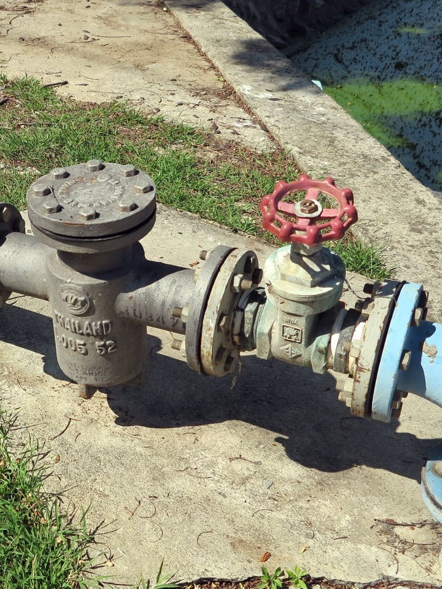 Metal pipes with valves, including one with a red handle, on concrete, outdoors.