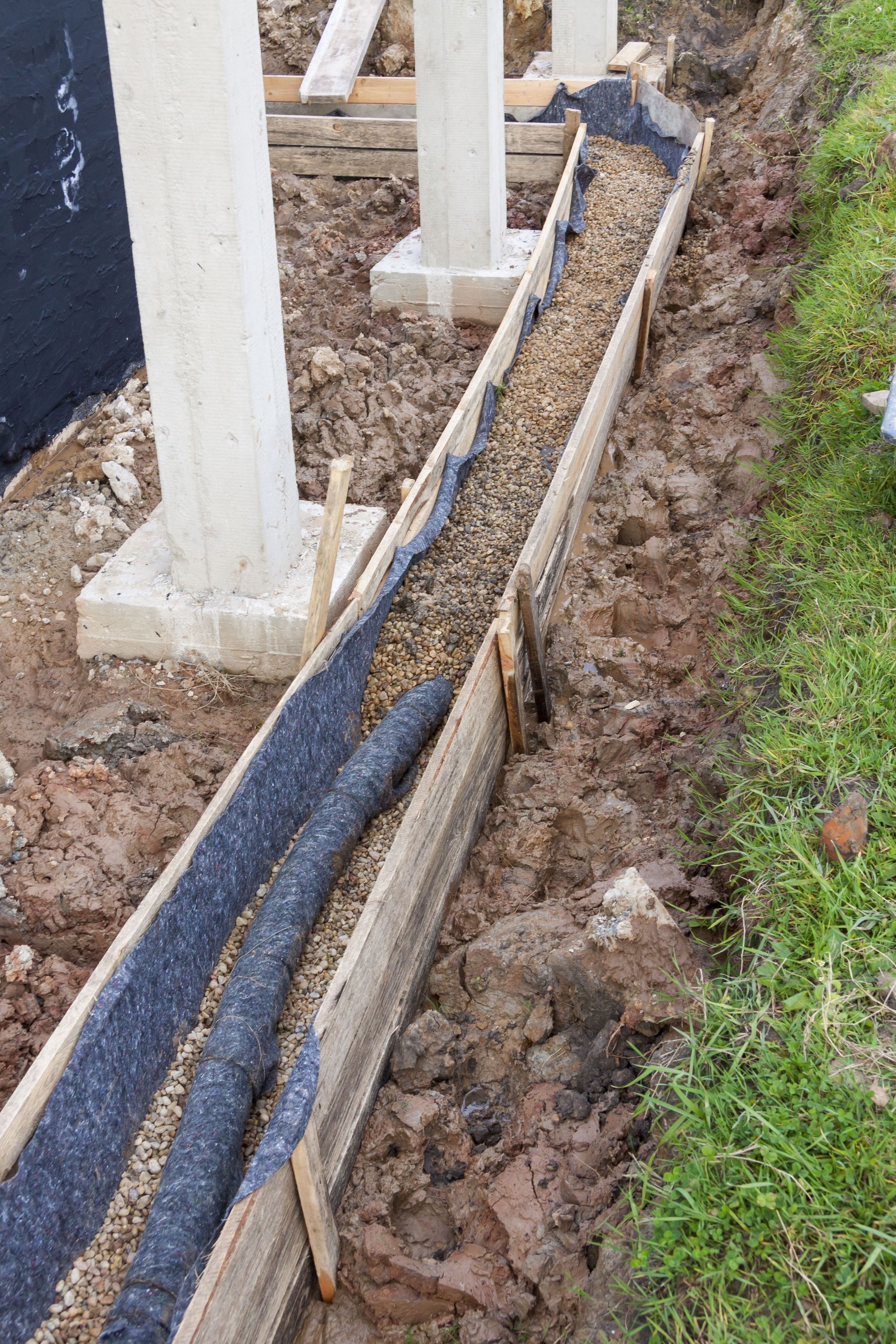 Construction site with a trench filled with gravel, lined with fabric, beside concrete pillars.