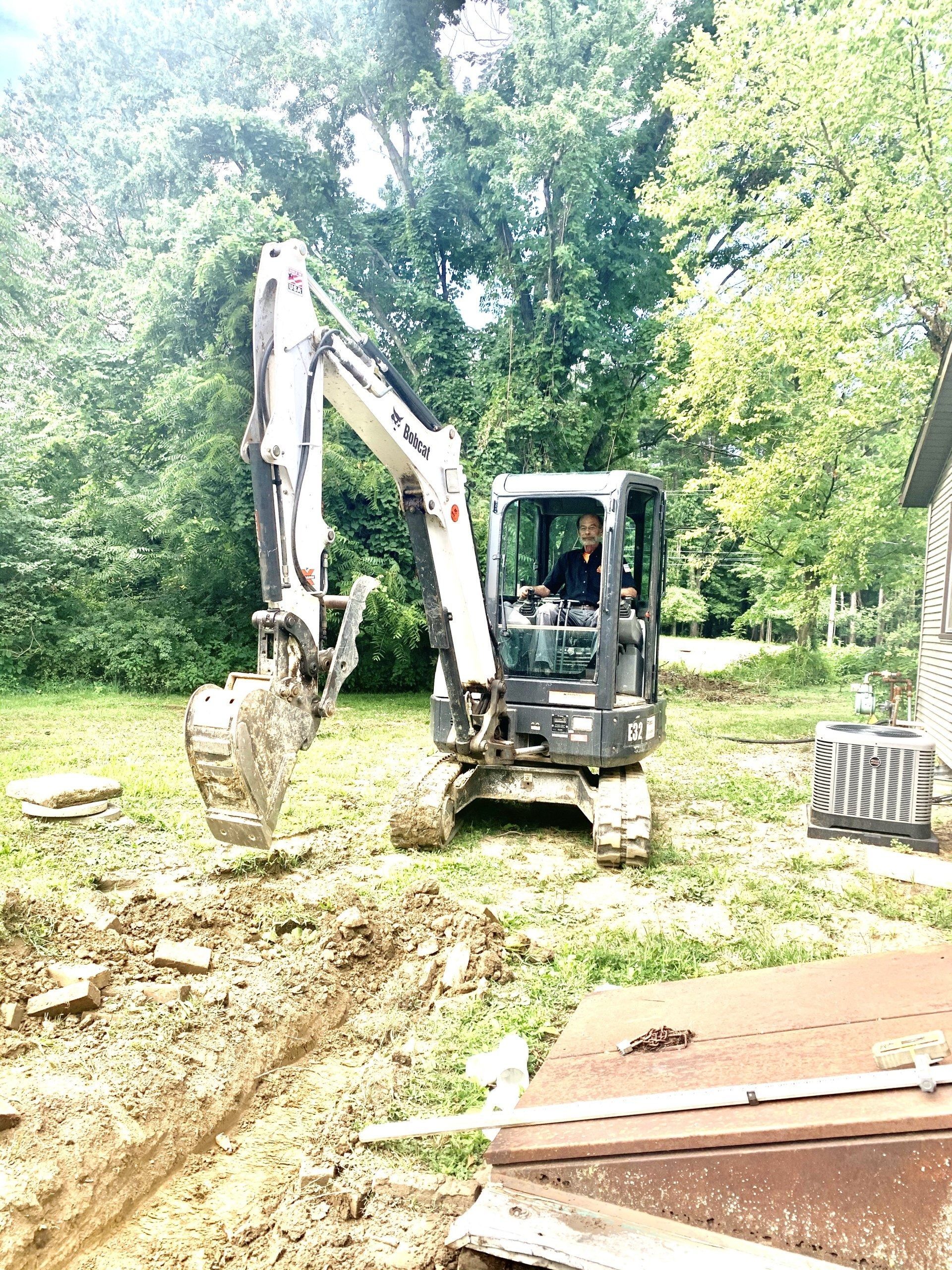 Person operating a small excavator in a grassy area near trees and a building.