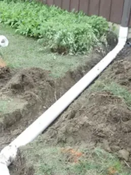 A white PVC drain pipe in a trench in a grassy yard, next to a brown fence and foliage.