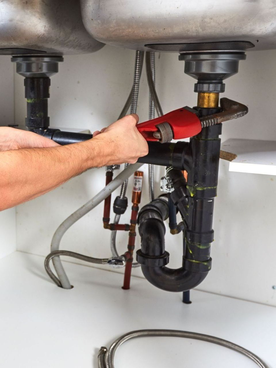 Plumber using a red wrench to repair a black pipe under a kitchen sink.