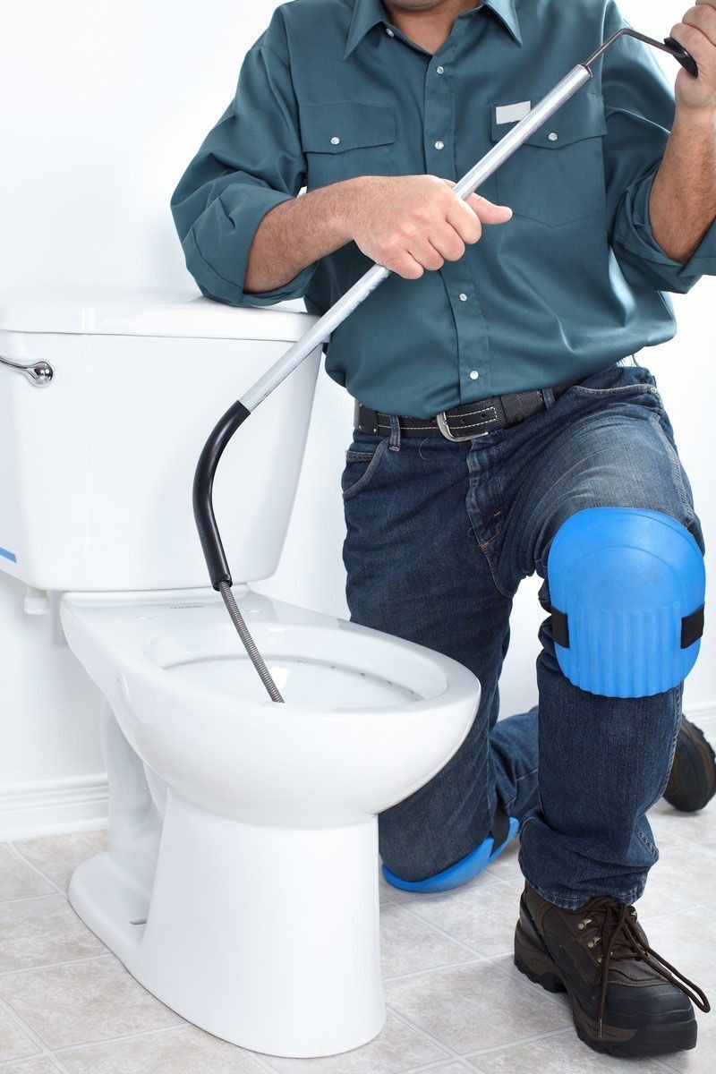 Plumber kneeling by a white toilet, using a plumbing snake. He wears blue knee pads, jeans, and a green shirt.
