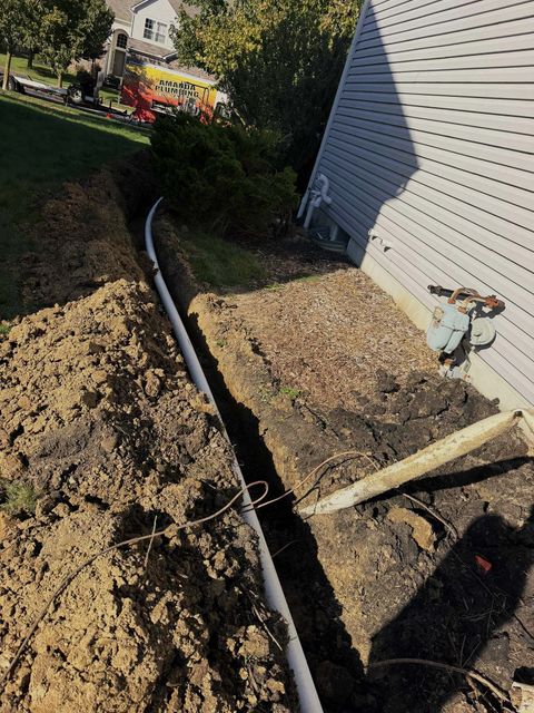 Trench dug beside a house, with black pipe laid in the dirt and tools on the ground.