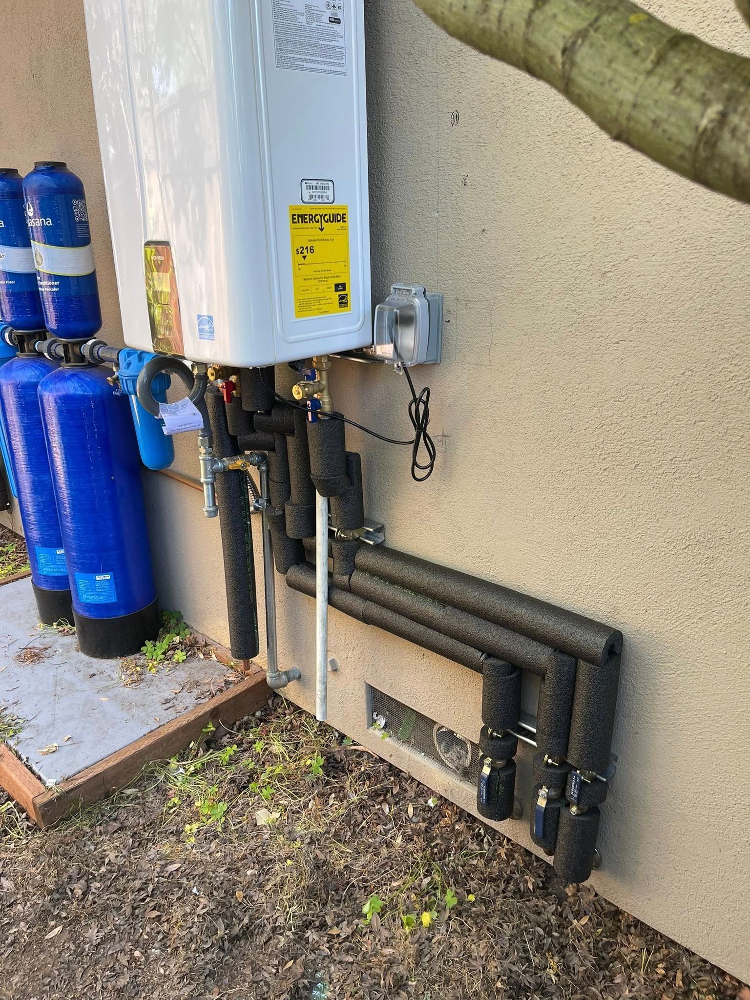 Outdoor water heater with blue filtration tanks and exposed pipes beside a stucco wall.