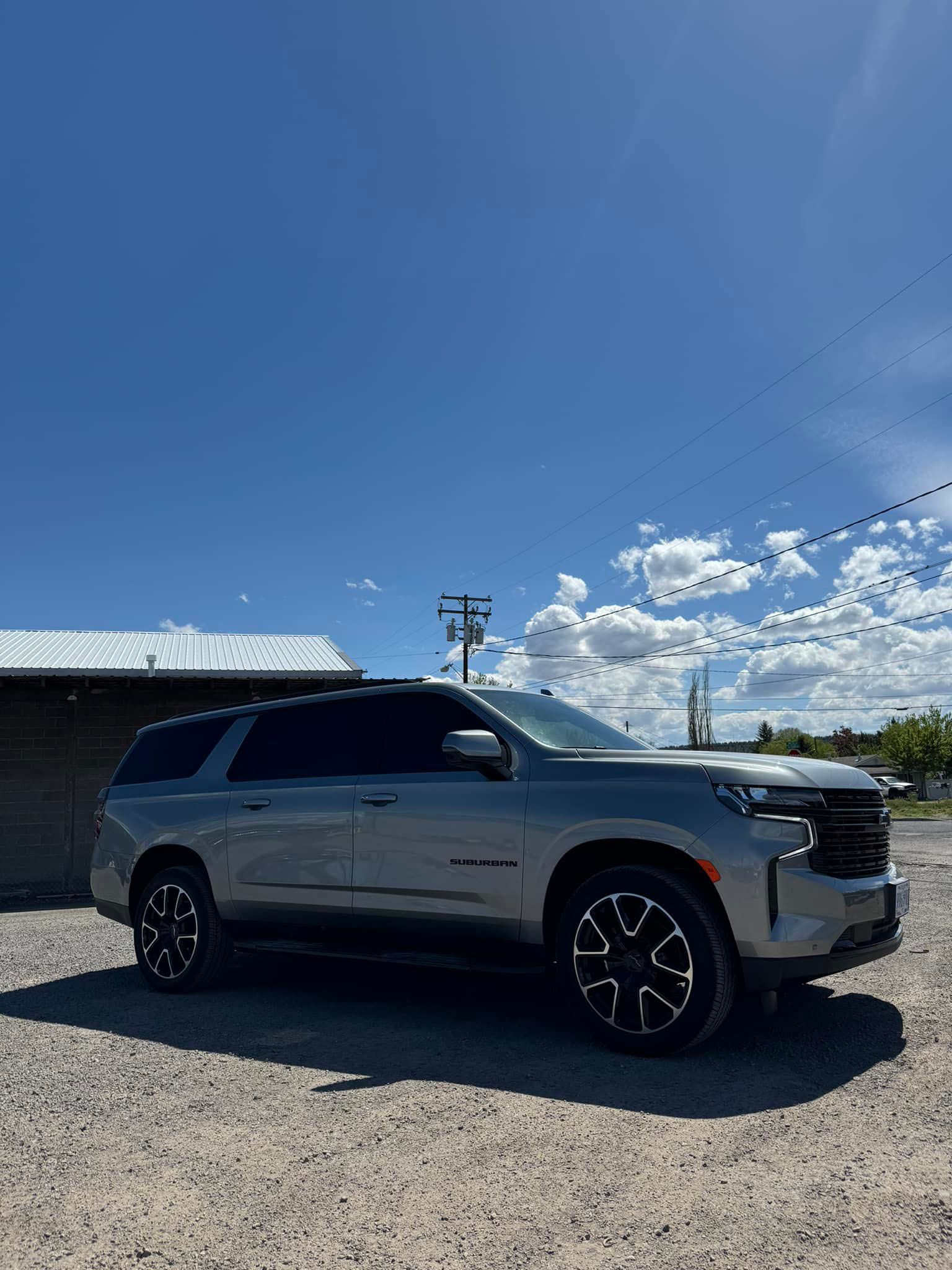 Gray Chevrolet Suburban SUV parked on gravel under a blue sky.
