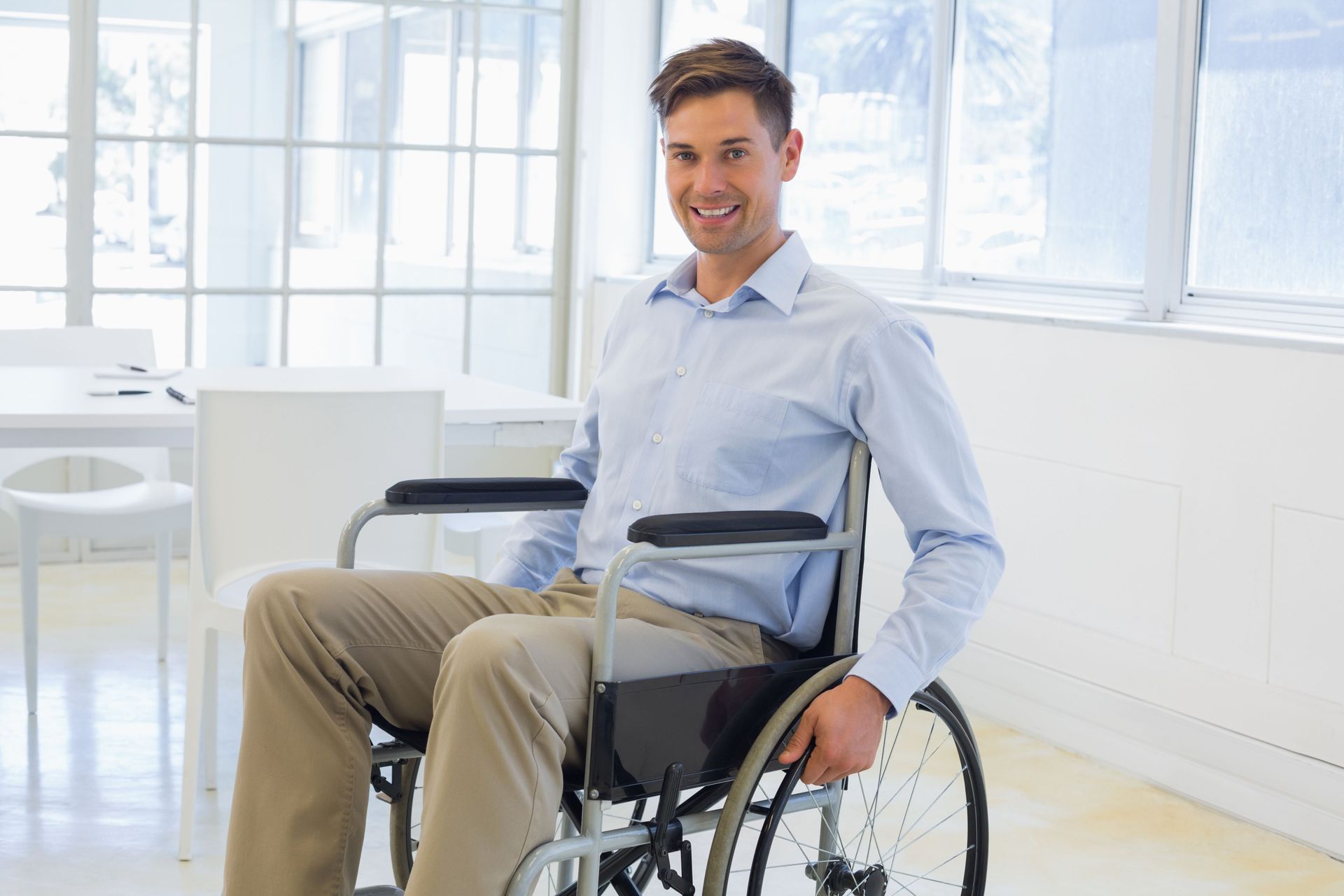 Man in wheelchair smiles in an office setting, wearing a blue shirt and khaki pants.