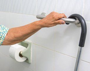 Person's hand holding a bathroom grab bar next to a toilet paper roll, white tiled wall.