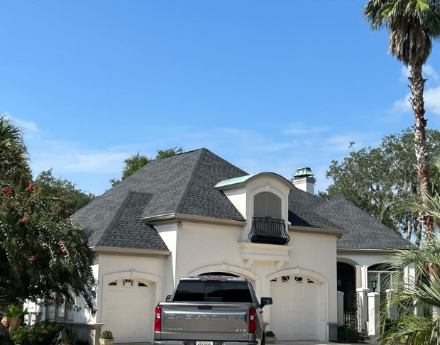 White stucco house with dark gray roof, two-car garage, and gray truck in driveway. Palm trees and blue sky.