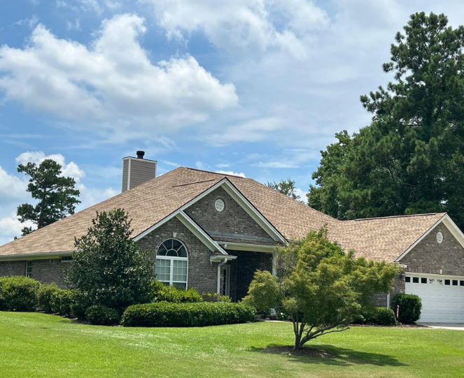 A brick house with a brown roof and chimney, surrounded by green trees and a lawn.