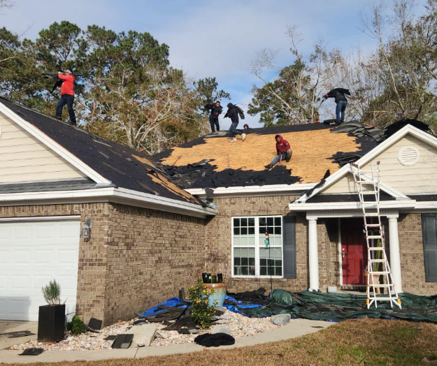 Roofing crew working on a house, removing old shingles. Brick exterior, ladder, tarp below.