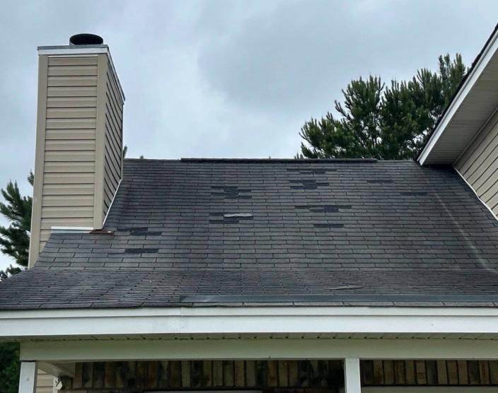 A residential roof with missing shingles next to a chimney and building siding, under an overcast sky.