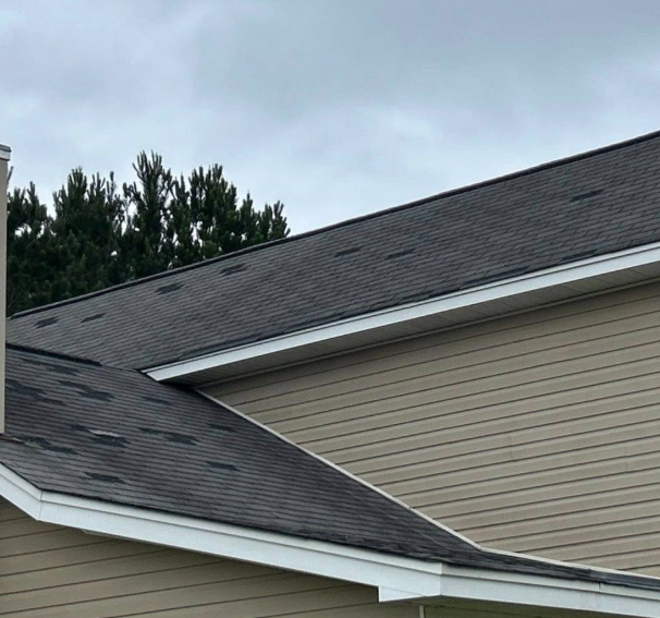 Gray shingled roof of a house with light tan siding against a cloudy sky.