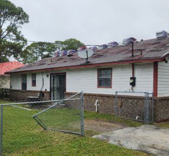 House with damaged roof and stacked shingles, chain-link fence, and overcast sky.