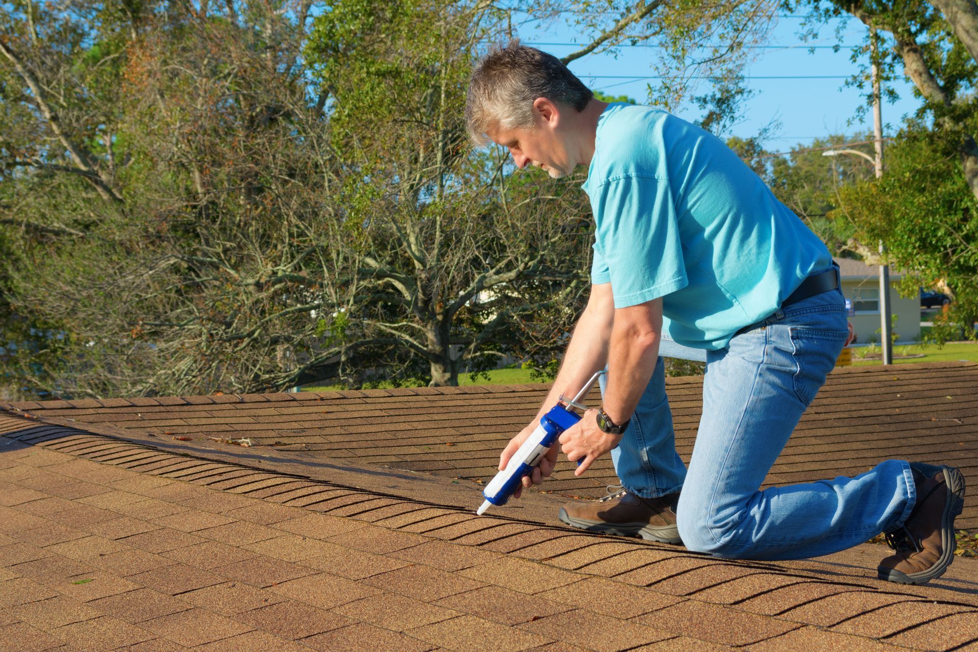 Man on a roof applying sealant with a caulk gun. Brown shingles and a sunny outdoor setting.